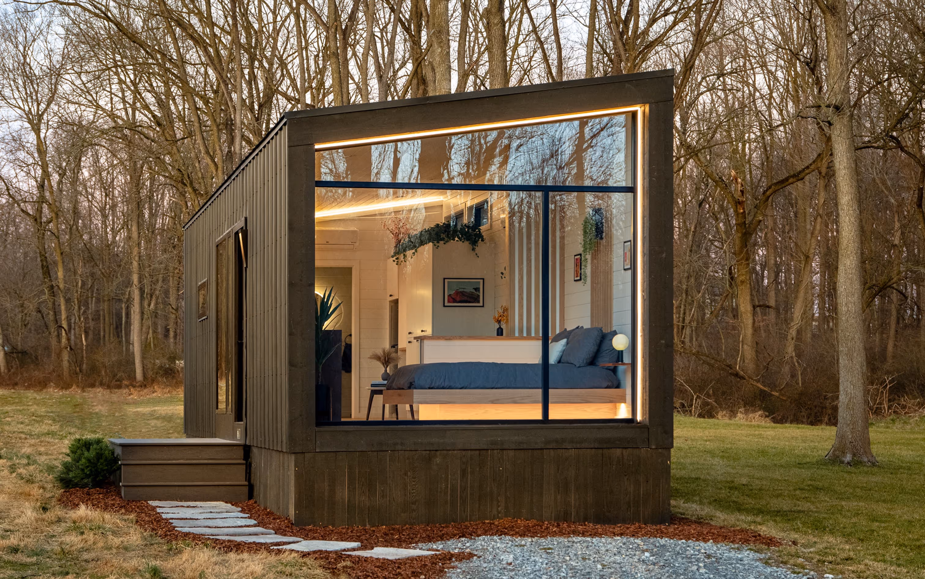 Modern tiny home with large window showing a cozy bedroom, surrounded by leafless trees and grass.