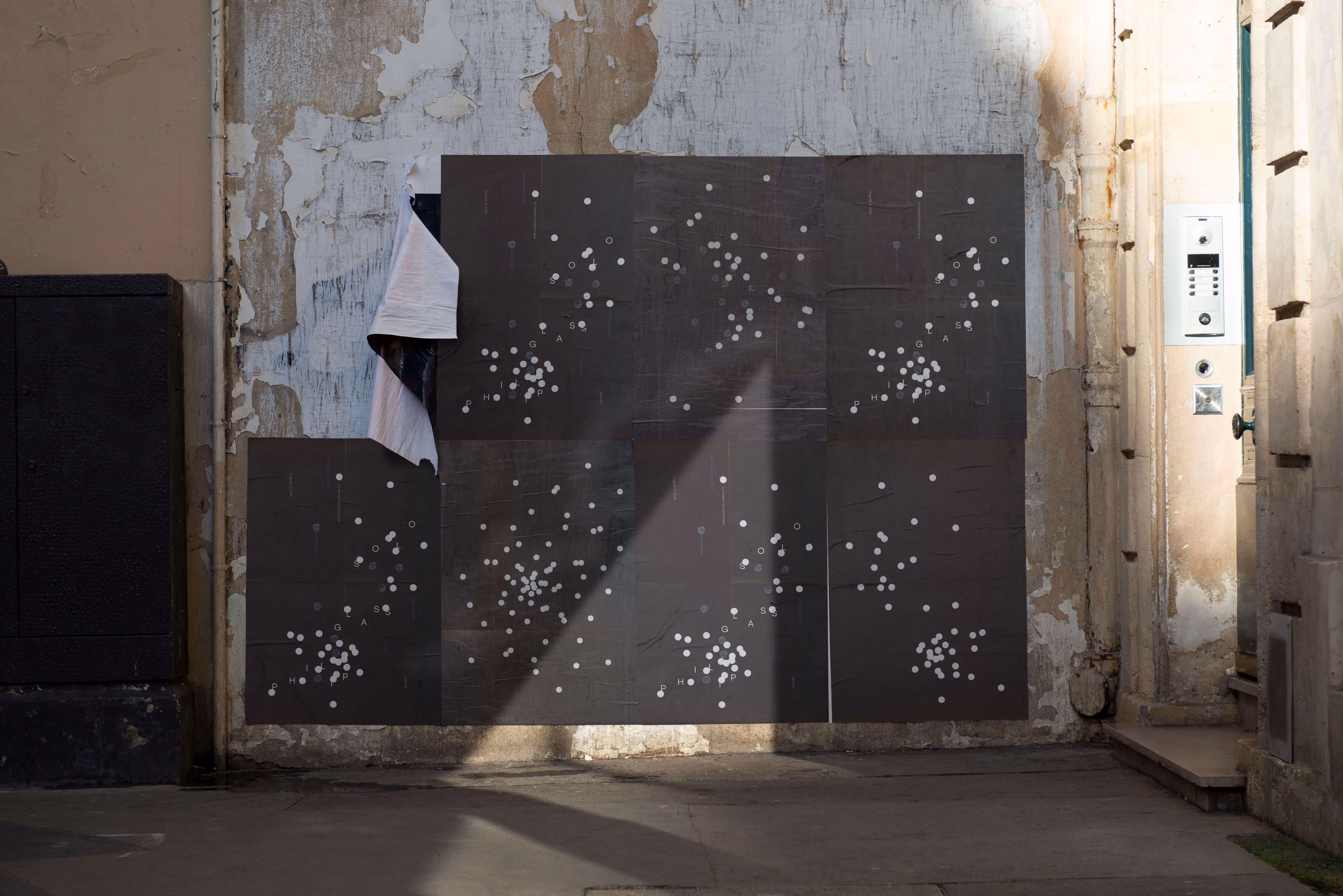 Black posters with white dots and letters pasted on a weathered wall, one corner peeling off.