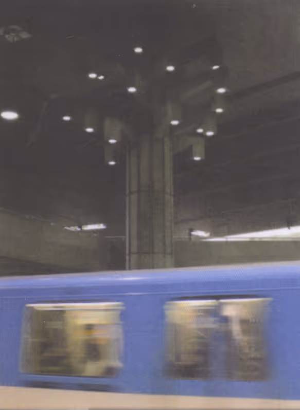 Blue metro train passing through a dimly lit concrete subway station with overhead lights.