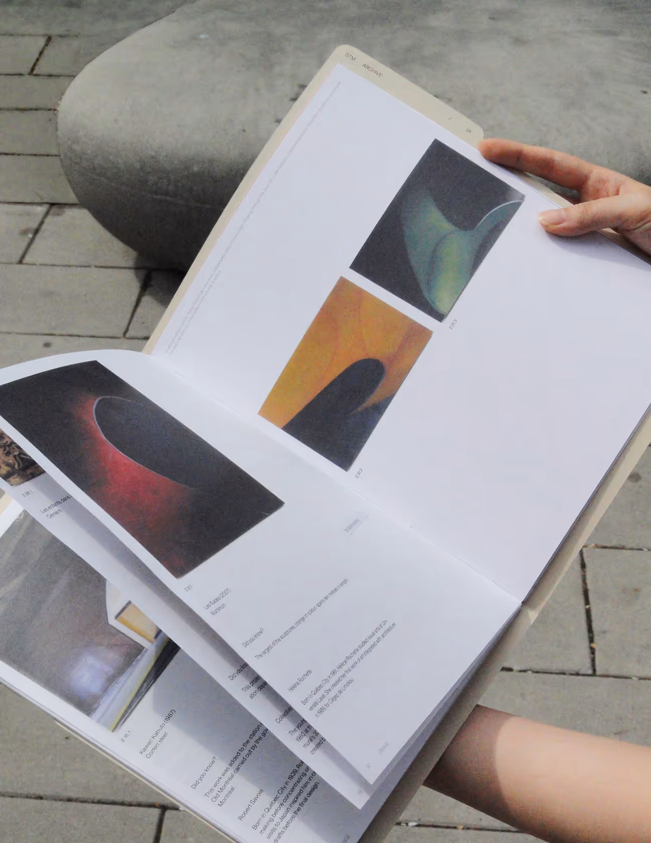 Hand holding an open art catalog showing abstract paintings on a stone bench background.