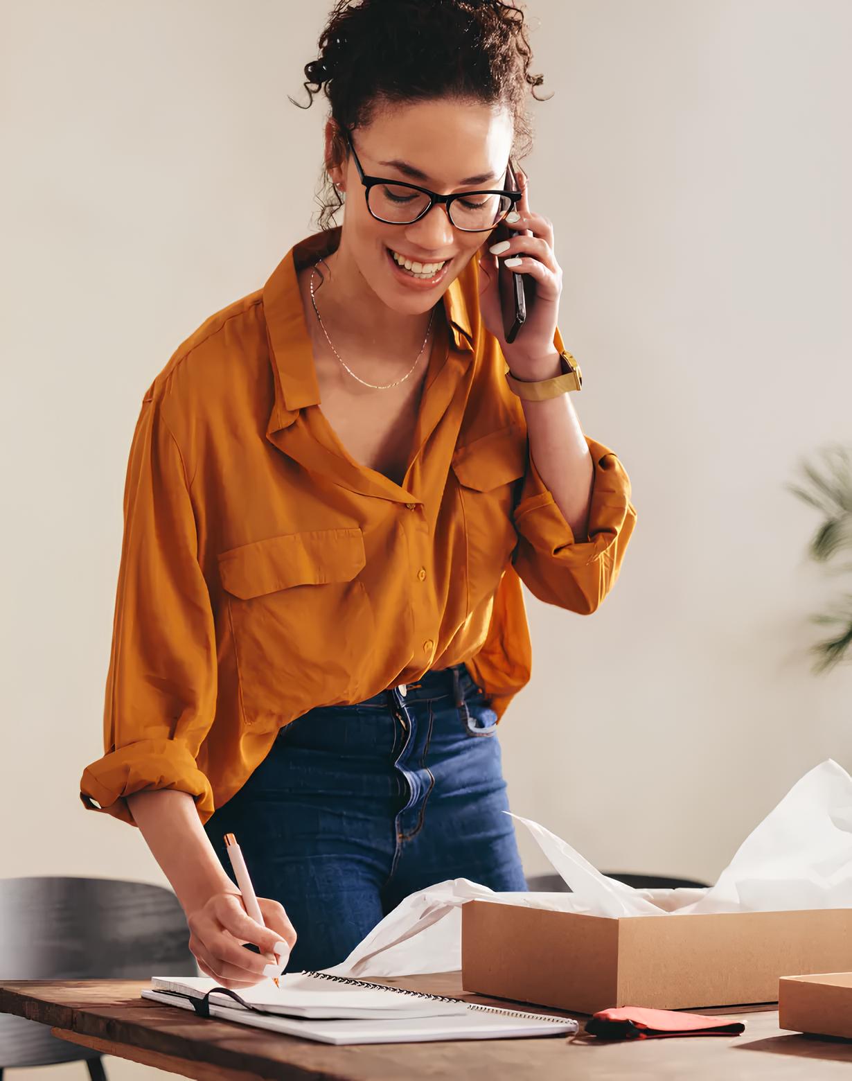 Smiling woman wearing glasses and an orange shirt talking on the phone while writing in a notebook at a wooden table.