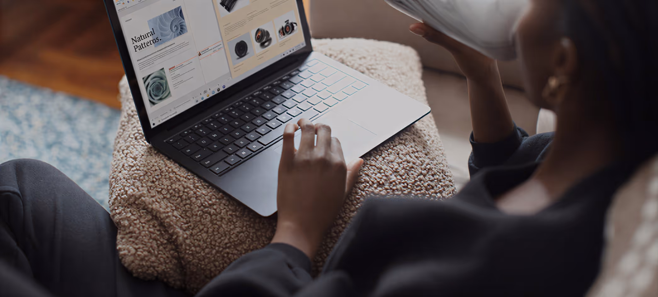Person using a laptop on a cushion with a webpage about natural patterns open, holding a cup in their other hand.