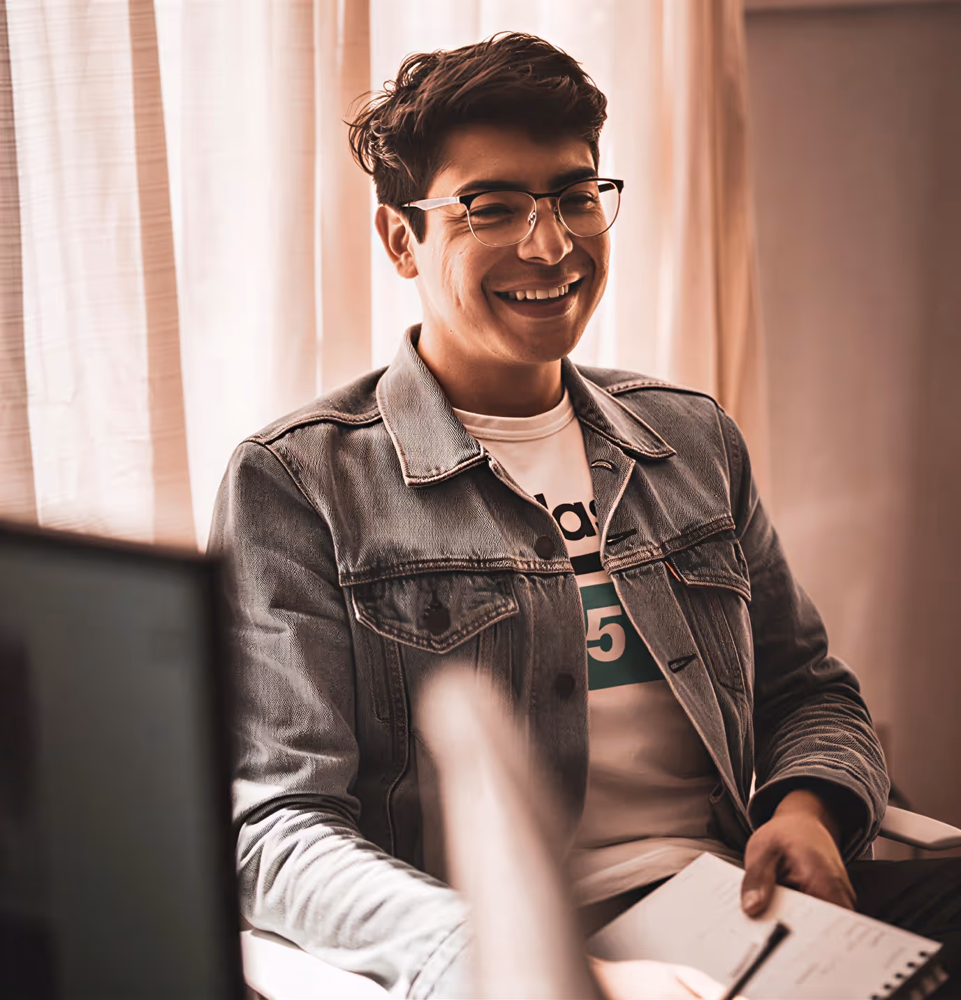 Smiling man with glasses wearing a denim jacket and holding a notebook and pen indoors.
