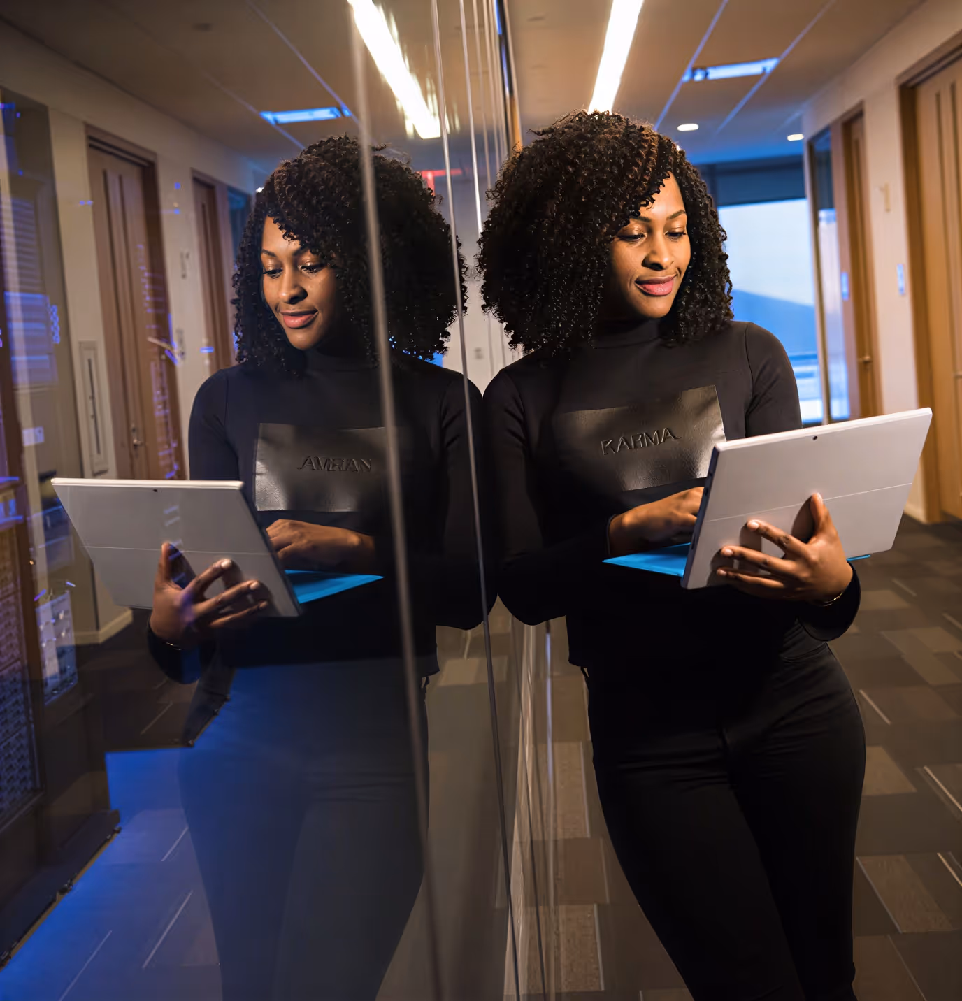 Woman with curly hair wearing black, holding a laptop and leaning against a glass wall in an office hallway.