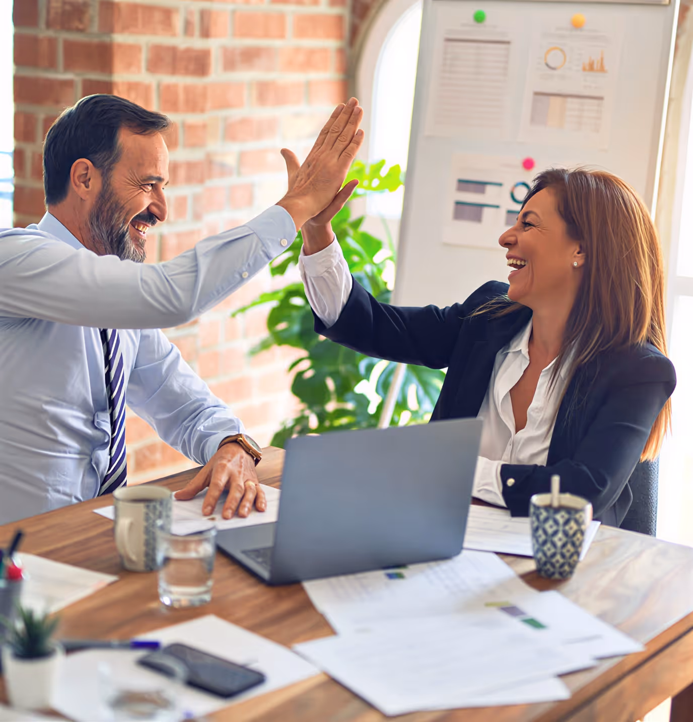 Two business professionals smiling and giving a high-five across a desk with a laptop and documents.