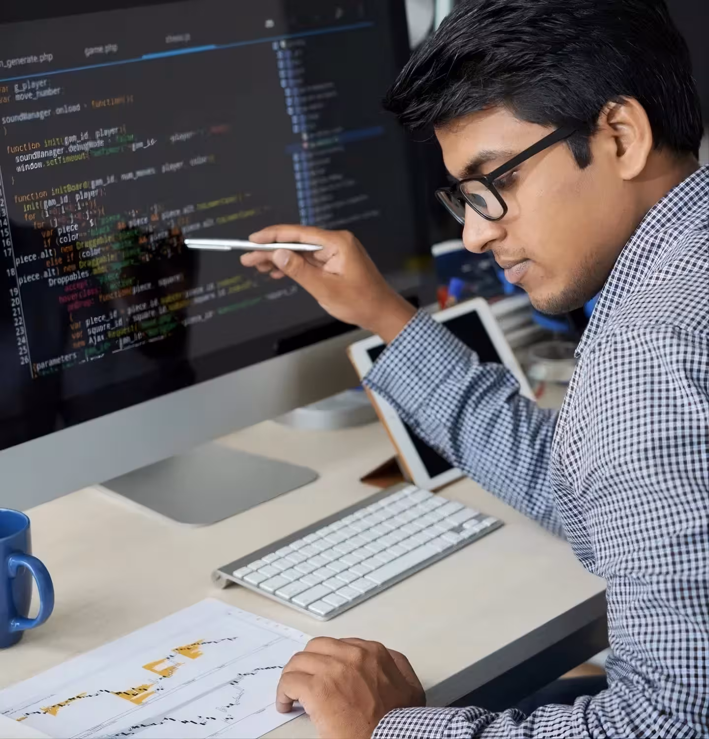 Man wearing glasses pointing at computer screen with code while viewing printed charts on desk.