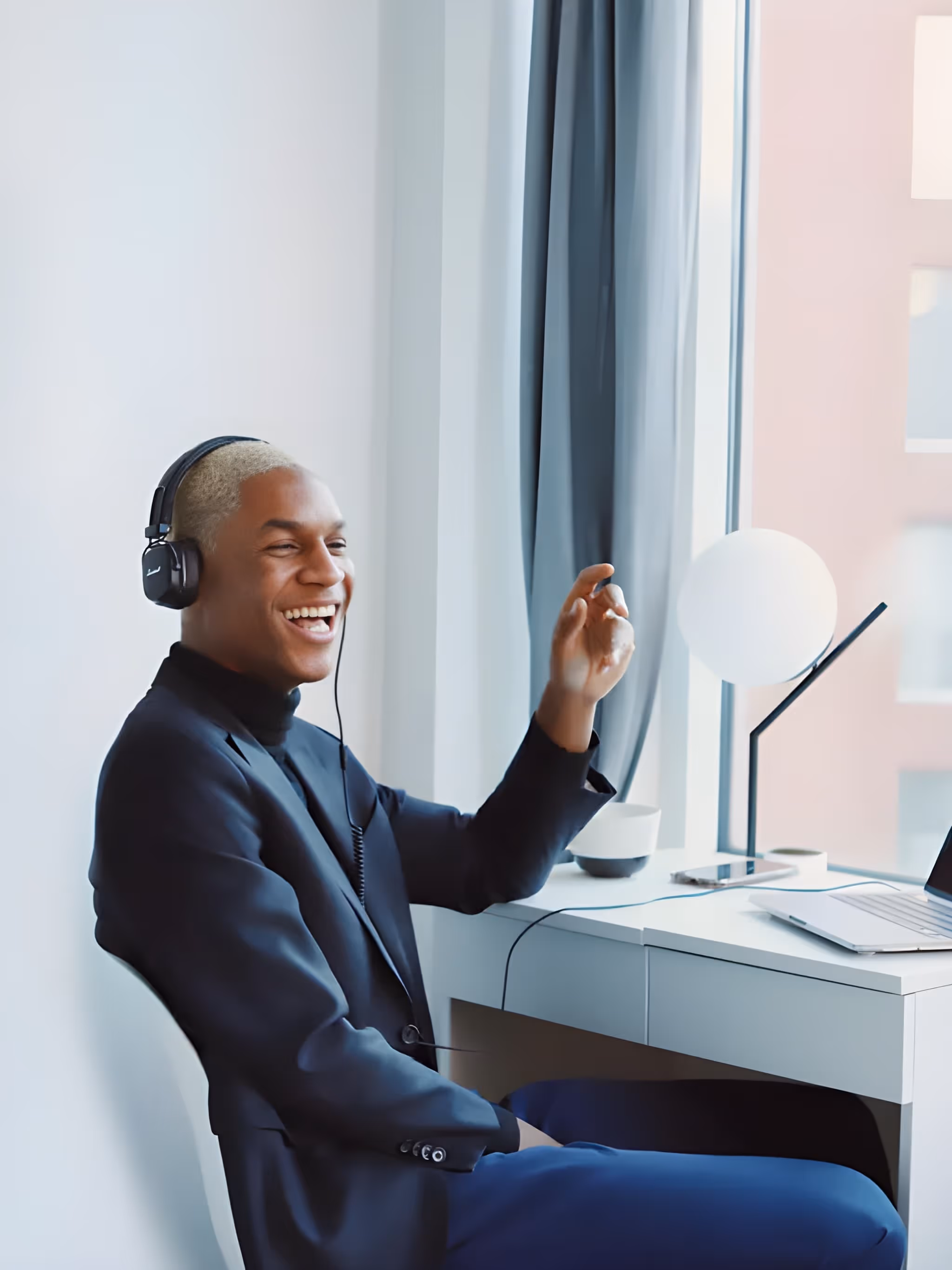 Smiling person with blonde hair wearing headphones and a dark blazer sitting at a desk with a laptop and lamp near a window.