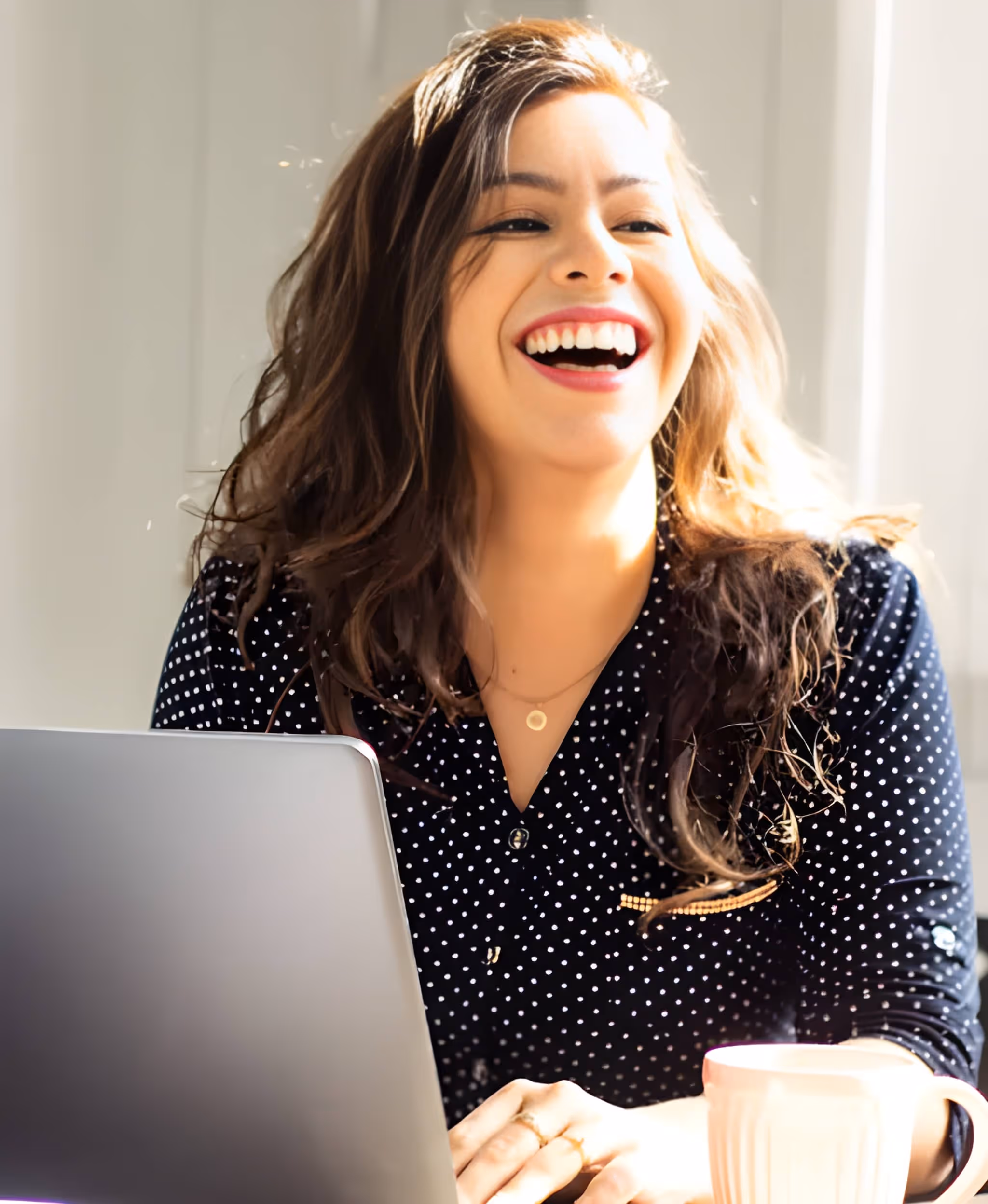 Happy woman with long brown hair laughing while sitting at a desk with a laptop and a white mug.