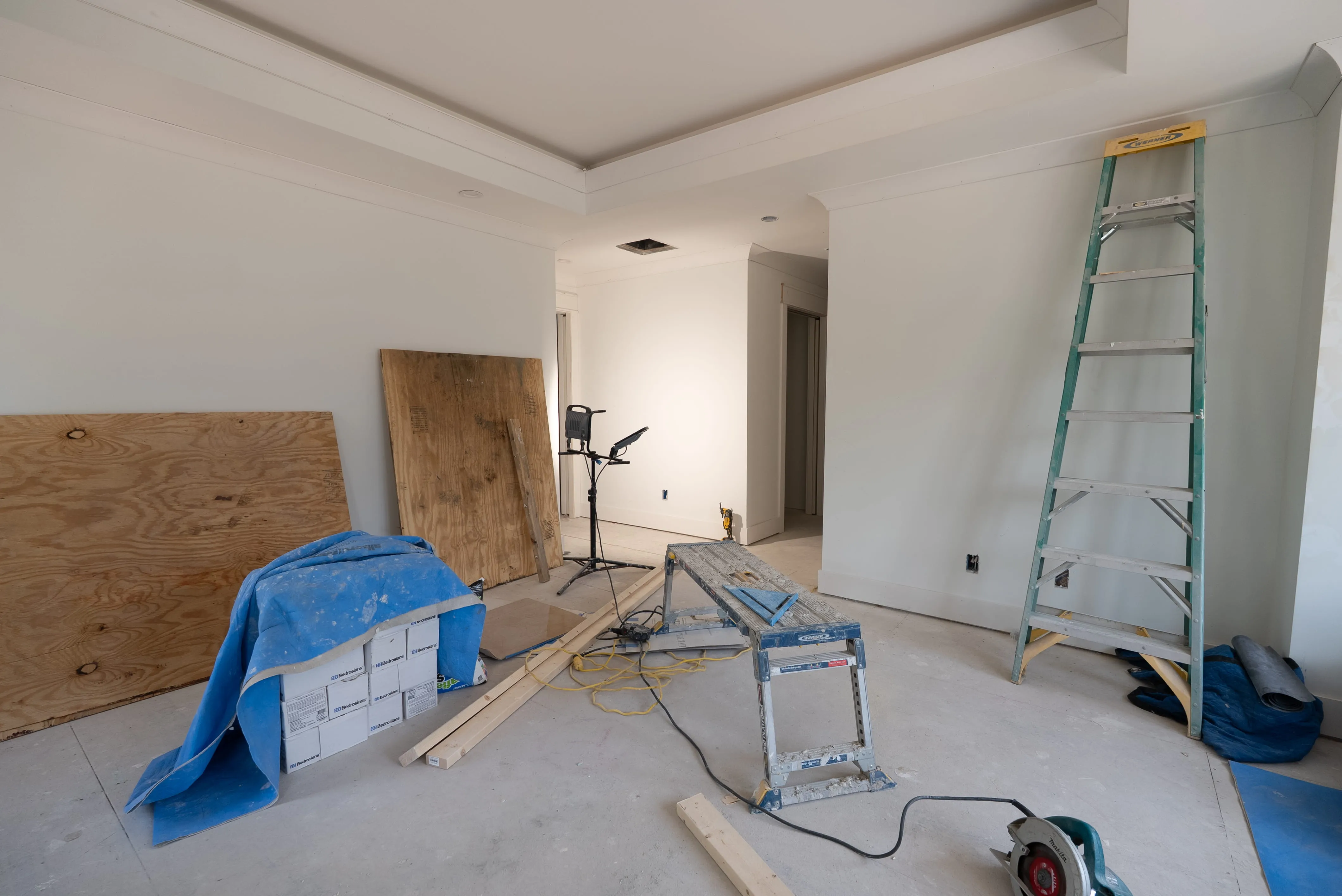 Interior room under renovation with ladder, workbench, circular saw, plywood, and electrical cords.