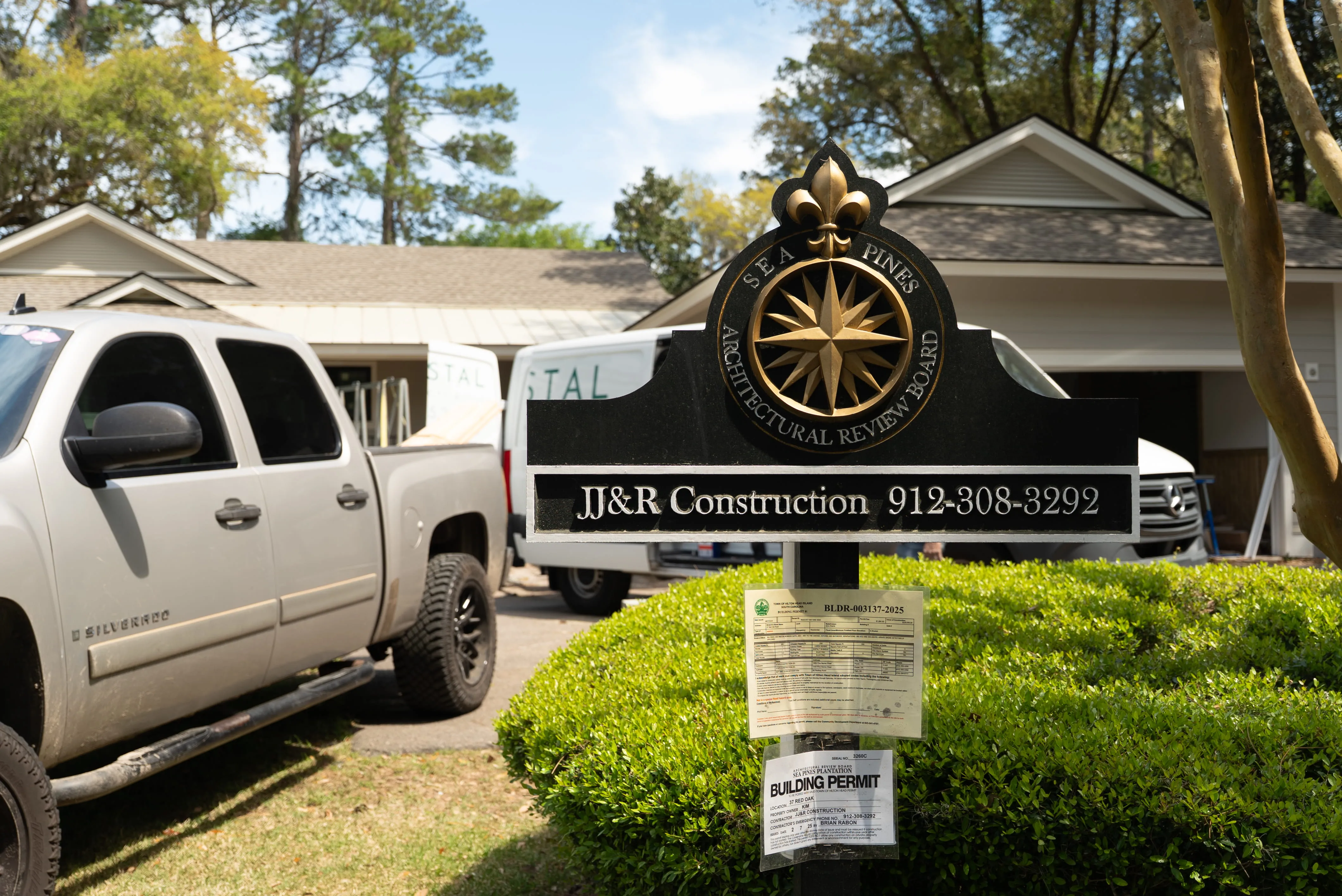 Sea Pines Architectural Review Board sign for JJ&R Construction with phone number and building permits on post near trucks.