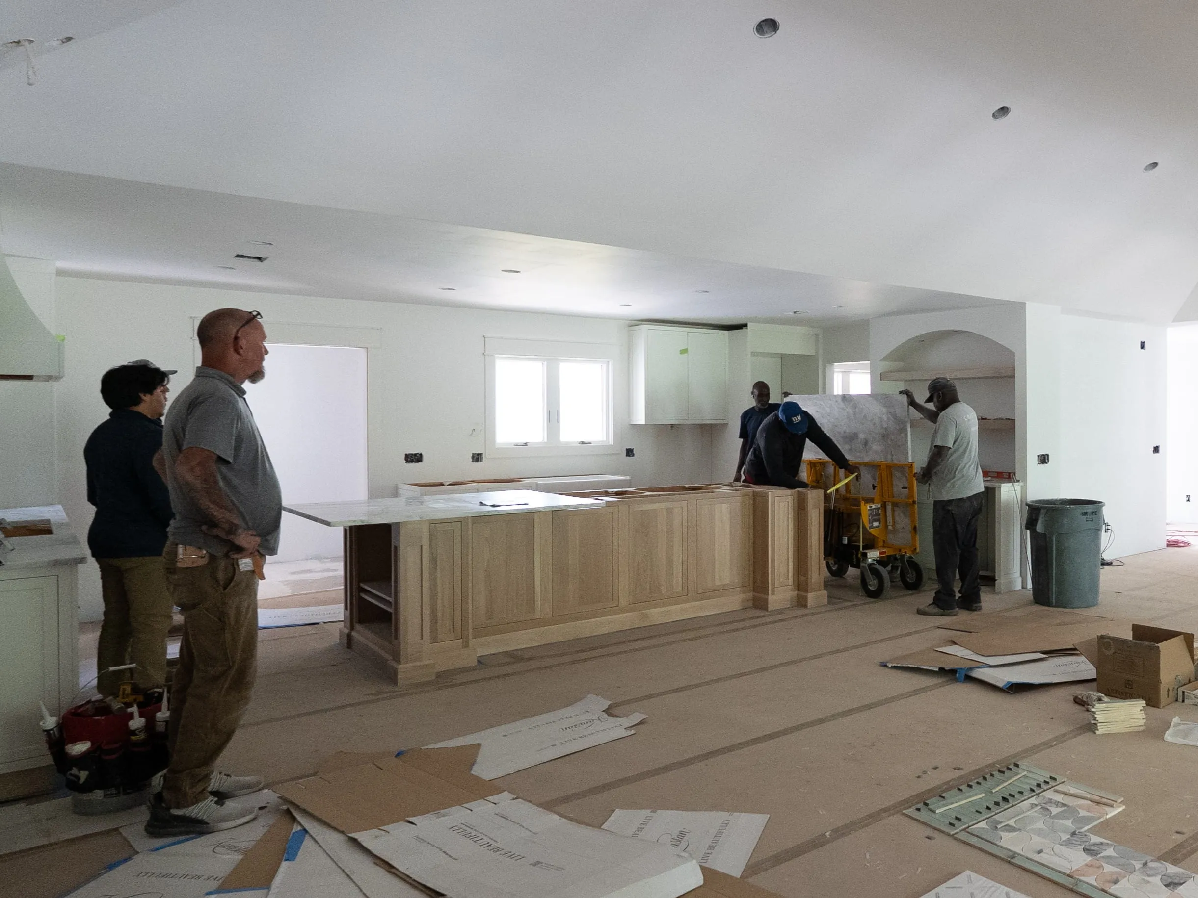Four workers installing a kitchen island with wood cabinetry and marble countertop in an unfinished room.