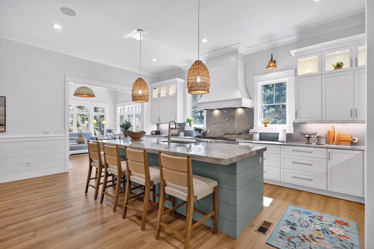 Modern kitchen with white cabinets, a large marble island with wood chairs, two wicker pendant lights, and hardwood floor.