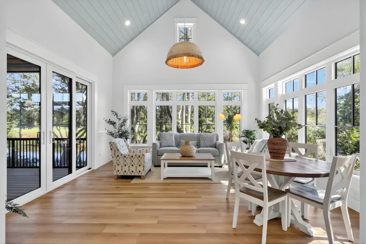 Bright living room with vaulted blue ceiling, large windows, gray sofa, armchair, wooden dining table, and indoor plants.
