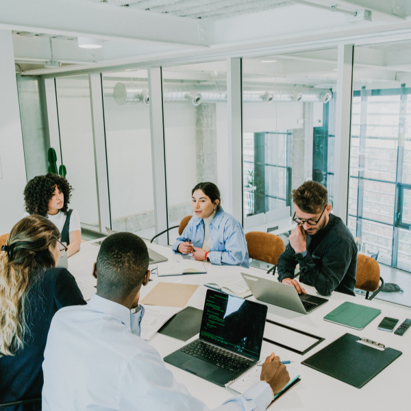 Group of five diverse people in a modern office meeting around a table with laptops and notebooks.