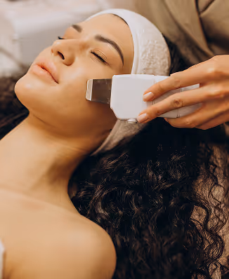 Woman with eyes closed receiving facial treatment with a skin scrubber device on her cheek.
