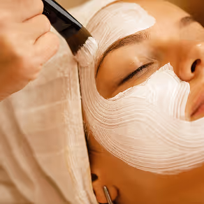 Person receiving a white facial mask application with a brush while wrapped in a towel.
