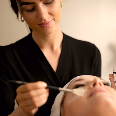 A beautician gently applies a facial mask with a brush to a relaxed client lying down with a towel wrapped around their head.