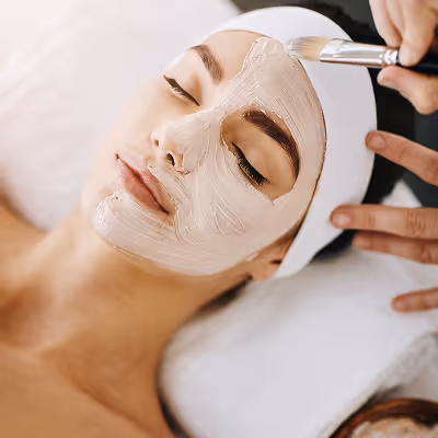 Woman with eyes closed receiving a creamy facial mask treatment applied with a brush at a spa.
