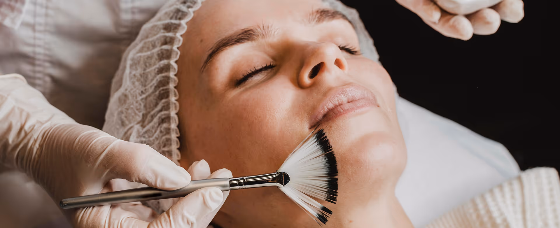 Close-up of a woman with eyes closed receiving a facial treatment with a fan brush by gloved hands.