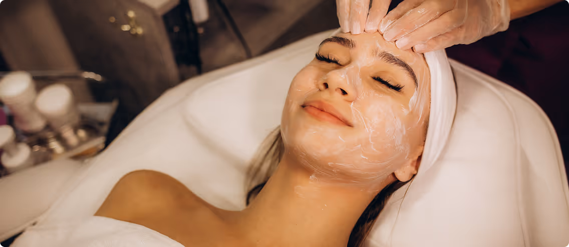 Smiling woman with eyes closed receiving a facial treatment with cream applied.