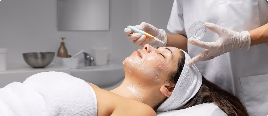 A woman lying down with eyes closed receiving a facial treatment where a skincare professional applies a cream using a brush on her face in a spa setting.