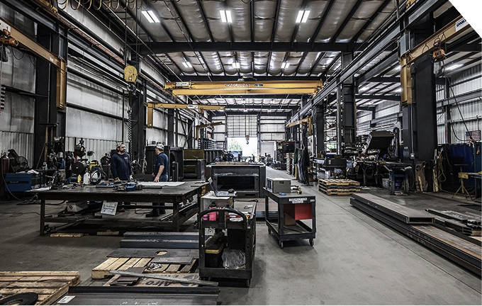 Spacious industrial workshop with metal beams, machinery, and two workers conversing near a large table.