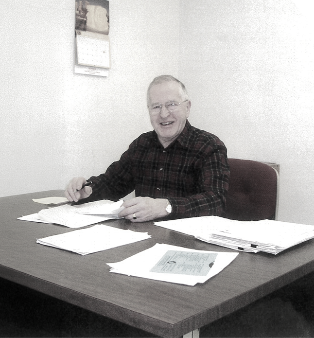 Smiling man in glasses and checkered shirt sitting at desk with papers and pen in an office room.
