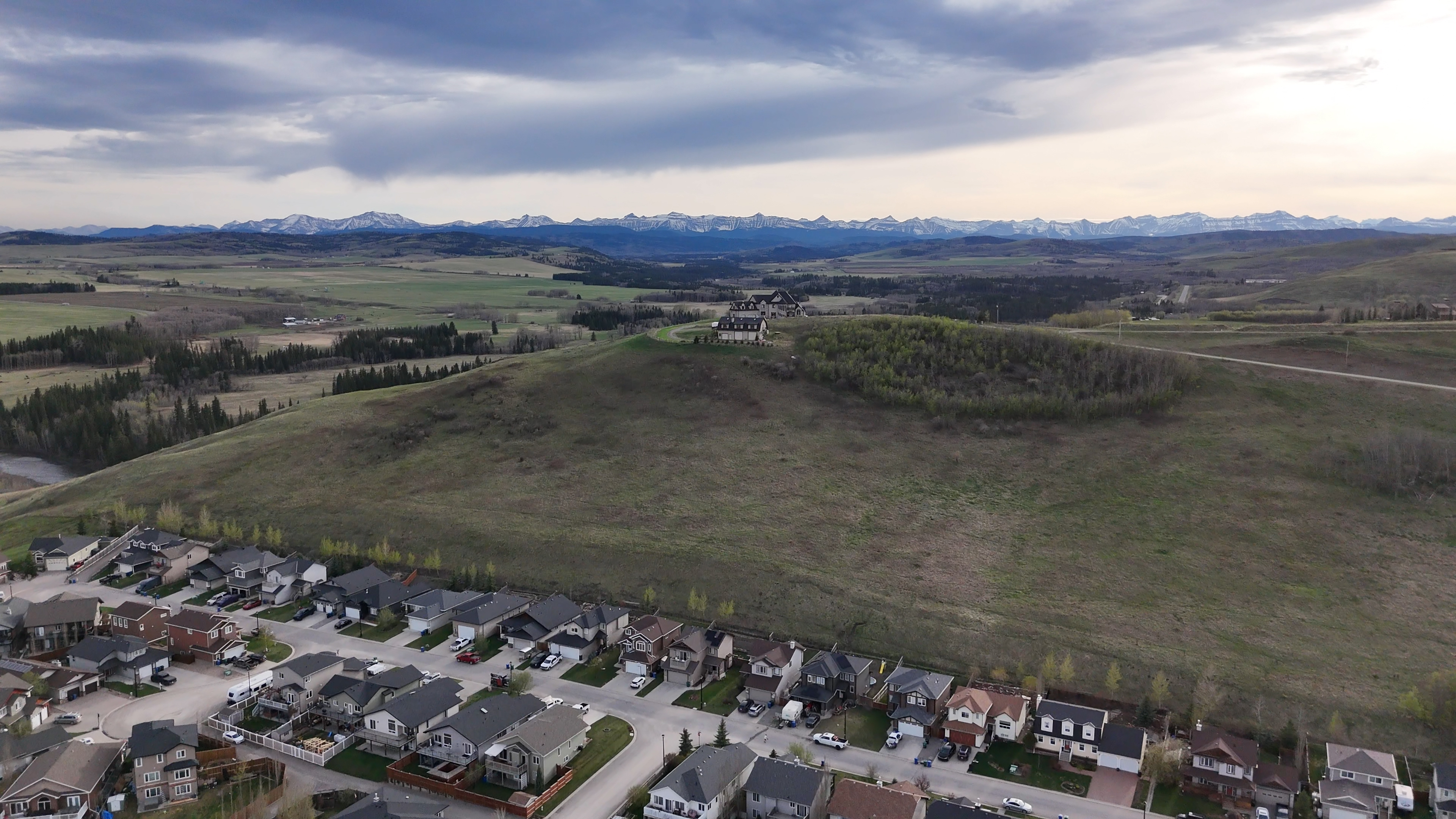 Aerial view of houses near a large grassy hill with scattered trees and distant snow-capped mountains.