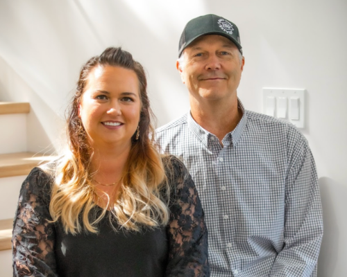 Smiling woman with long blond hair and man in checkered shirt and cap sitting indoors by stairs.