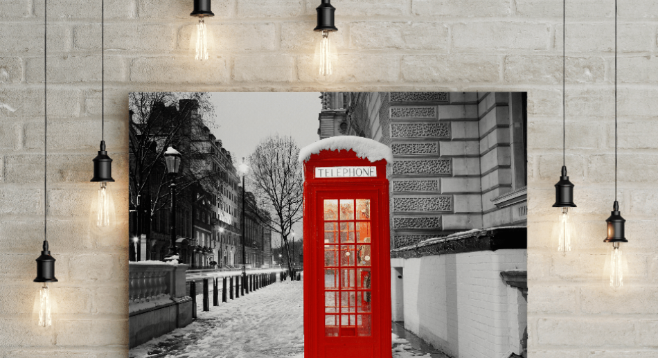 Wall-mounted black and white photo of a snowy street featuring a bright red telephone booth with snow on top, surrounded by hanging light bulbs.