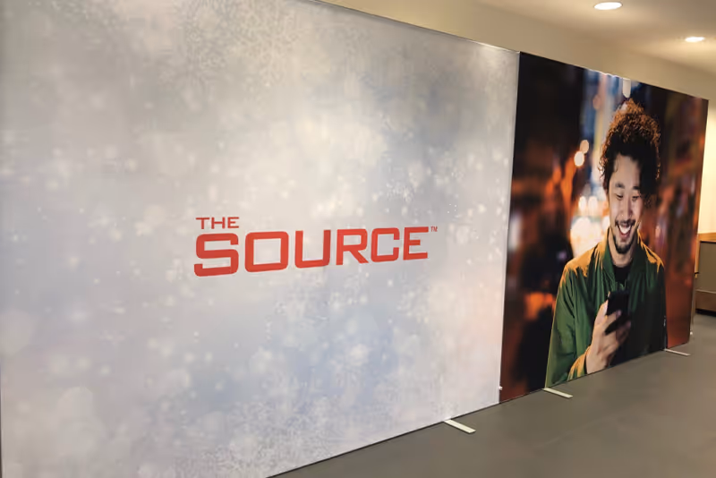 Large display panels with The Source logo on a white background and a smiling man looking at his phone.