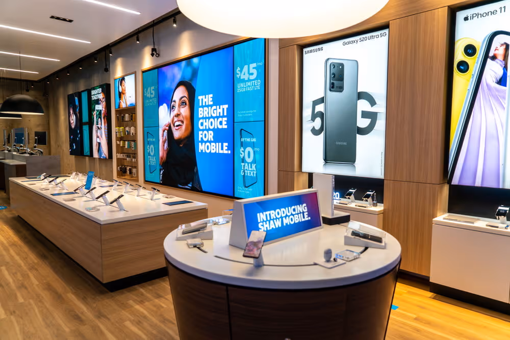 Interior of a mobile phone store with wooden displays featuring Samsung Galaxy S20 Ultra 5G, iPhone 11, and a sign introducing Shaw Mobile.