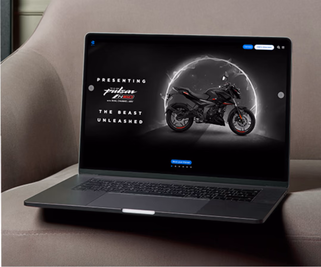 Laptop on a beige sofa displaying a black Pulsar N160 motorcycle with the text 'Presenting Pulsar N160 The Beast Unleashed' on screen.