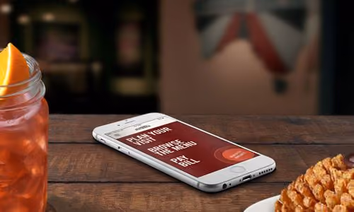 Smartphone on a wooden table displaying options to play music, browse menu, and pay bill in a restaurant setting with drink and food nearby.