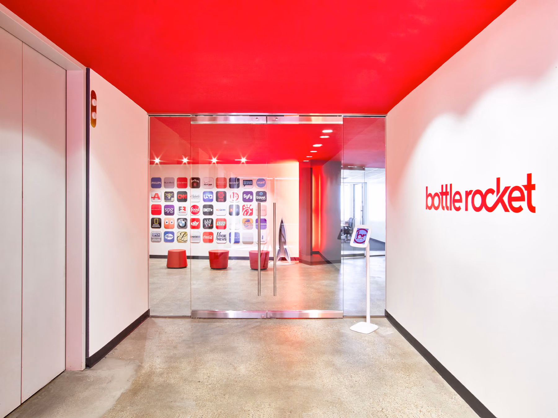 Modern office entrance with red ceiling, glass doors, red stools, a wall with various brand logos, and a 'bottle rocket' sign on the right wall.