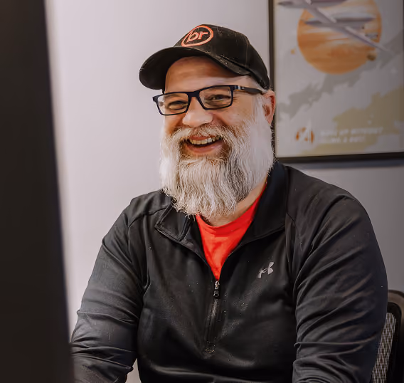 Smiling man with glasses, white beard, black cap, and black jacket sitting at a desk looking at a computer.