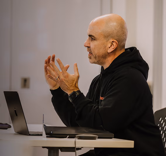 Man with a bald head and black hoodie speaking and gesturing with hands while sitting at a desk with a laptop and binder.