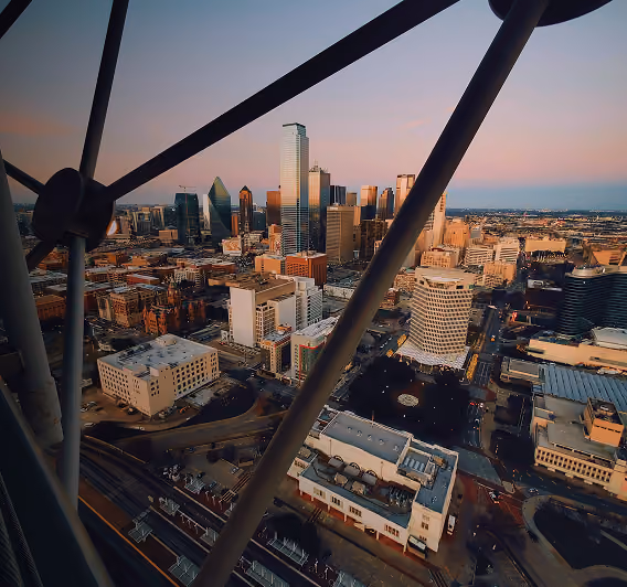 Aerial view of a city skyline at sunset with tall skyscrapers, framed partly by angled metal beams in the foreground.