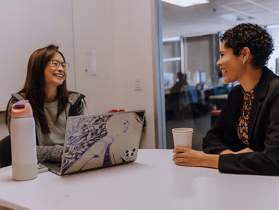 Two women smiling and talking across a table with a laptop and a coffee cup in an office.