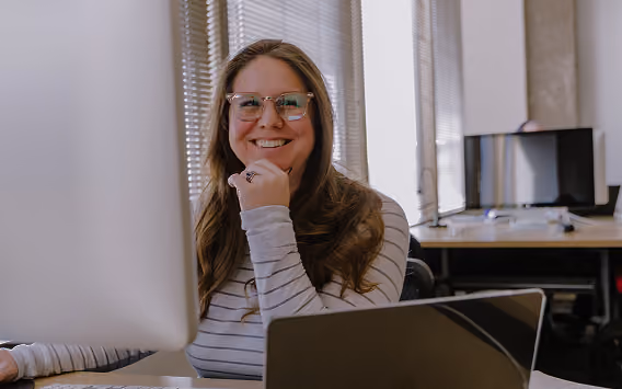 Smiling woman with glasses working at desk with computer monitors in a bright office.