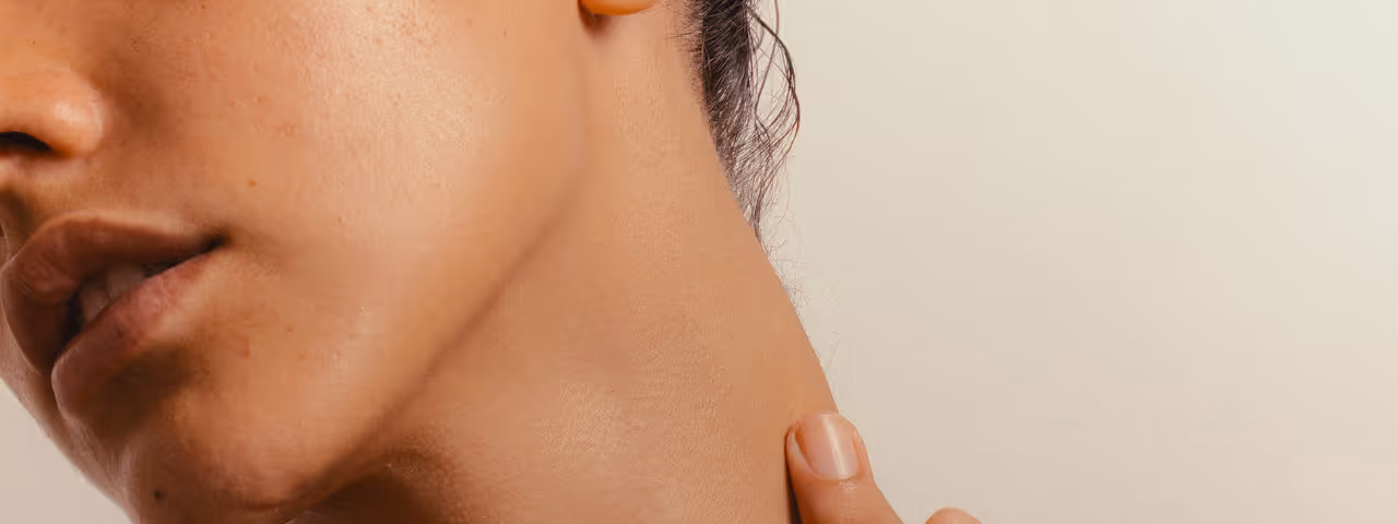Close-up of a person touching their neck with their hand, showing part of the face, jawline, and ear against a plain background.