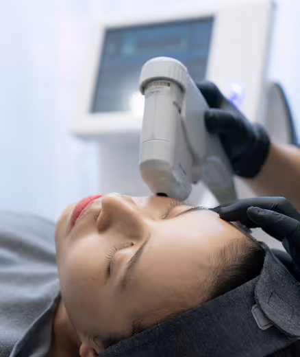 A woman receiving a cosmetic skin treatment on her forehead with a handheld device by a gloved professional.