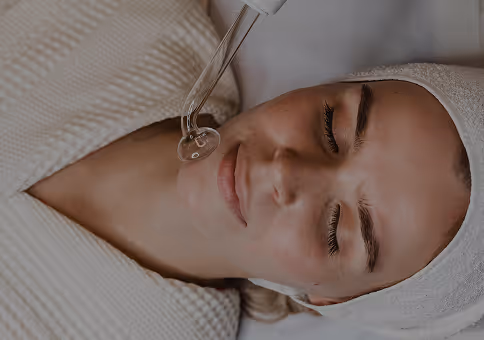 Woman with closed eyes receiving a facial serum application with a dropper while wearing a white towel headband and robe.
