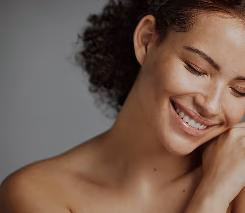 Smiling woman with closed eyes and curly hair resting her face gently on her hand against a gray background.