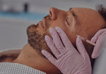 Man with closed eyes receiving a cosmetic injection in his temple from a gloved medical professional.