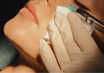 Close-up of a person receiving a cosmetic injection near the cheek, with gloved hands holding a syringe and cotton.