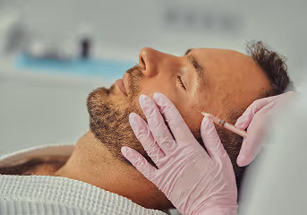 Close-up of a man receiving a cosmetic injection on his cheek while lying down with eyes closed.