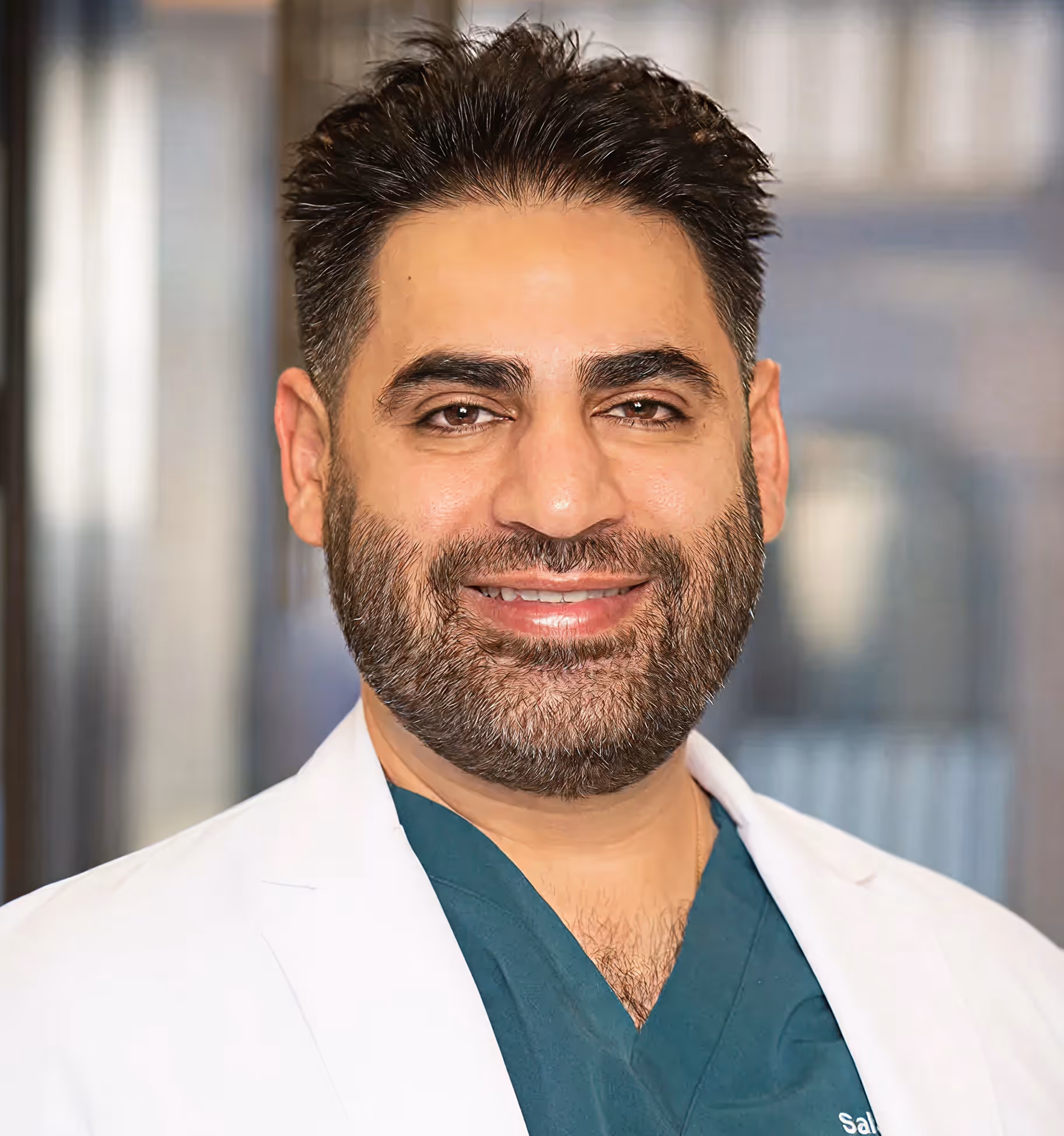 Smiling man with dark hair and beard wearing teal medical scrubs and a white lab coat.