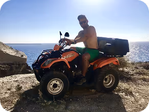 Man wearing sunglasses and green shorts sitting on an orange all-terrain vehicle near a rocky coastline with the ocean in the background.