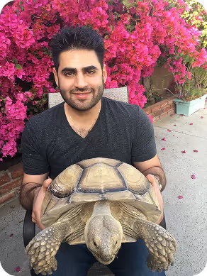 Man with dark hair and beard sitting outdoors holding a large tortoise in front of pink flowering bushes.